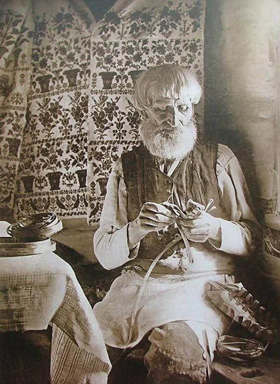 Photographie sépia : intérieur traditionnel, rushnyky accrochés au mur, homme au travail artisanal.