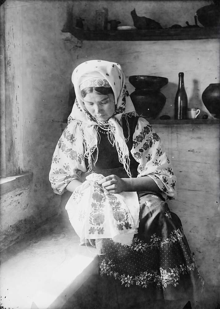 Femme en costume brodant un rushnyk près d’une fenêtre, intérieur rustique.